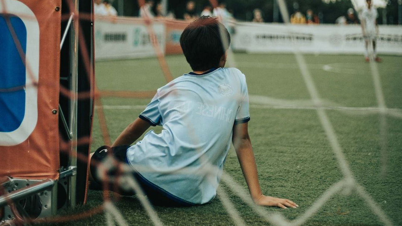 A young football player in a light blue kit sitting on the ground inside the goal area, looking out onto the pitch during a youth match.