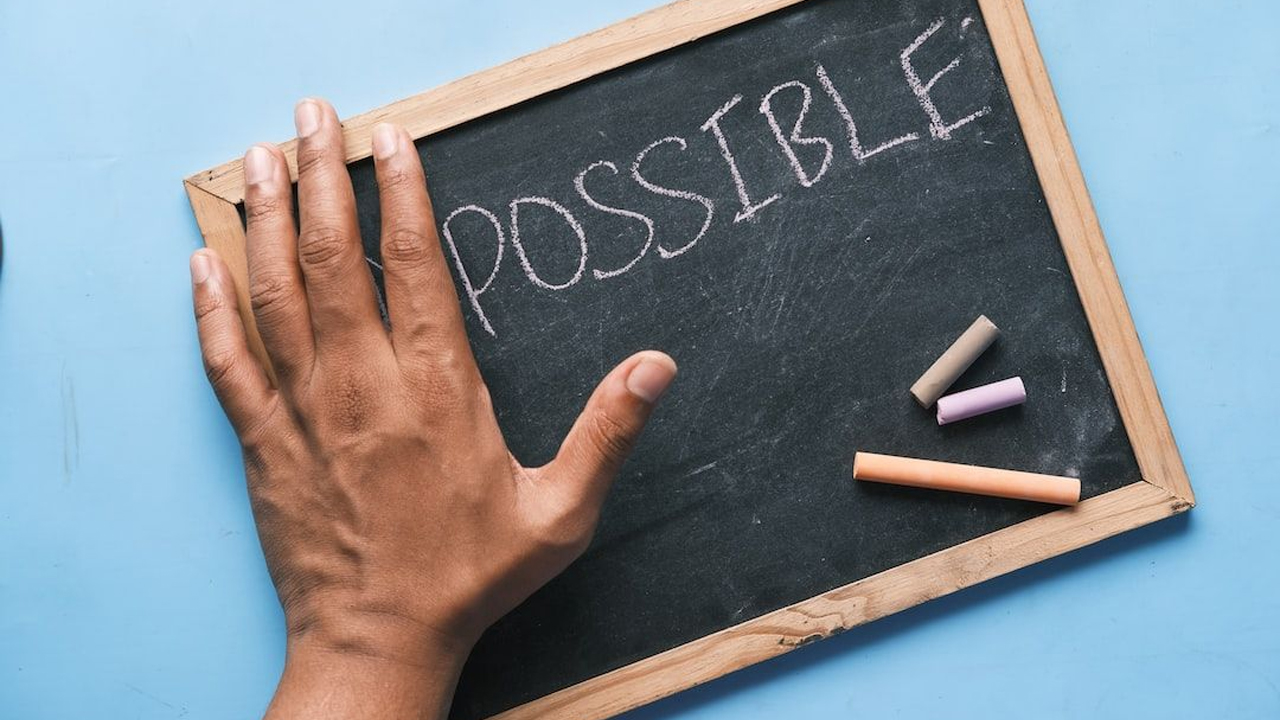A hand rests on a small chalkboard with the word “POSSIBLE” written on it in chalk, with pieces of coloured chalk beside it.