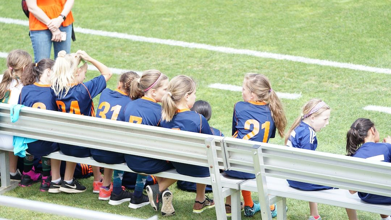 A girls’ youth football team sitting together on the subs bench, watching the game from the touchline.
