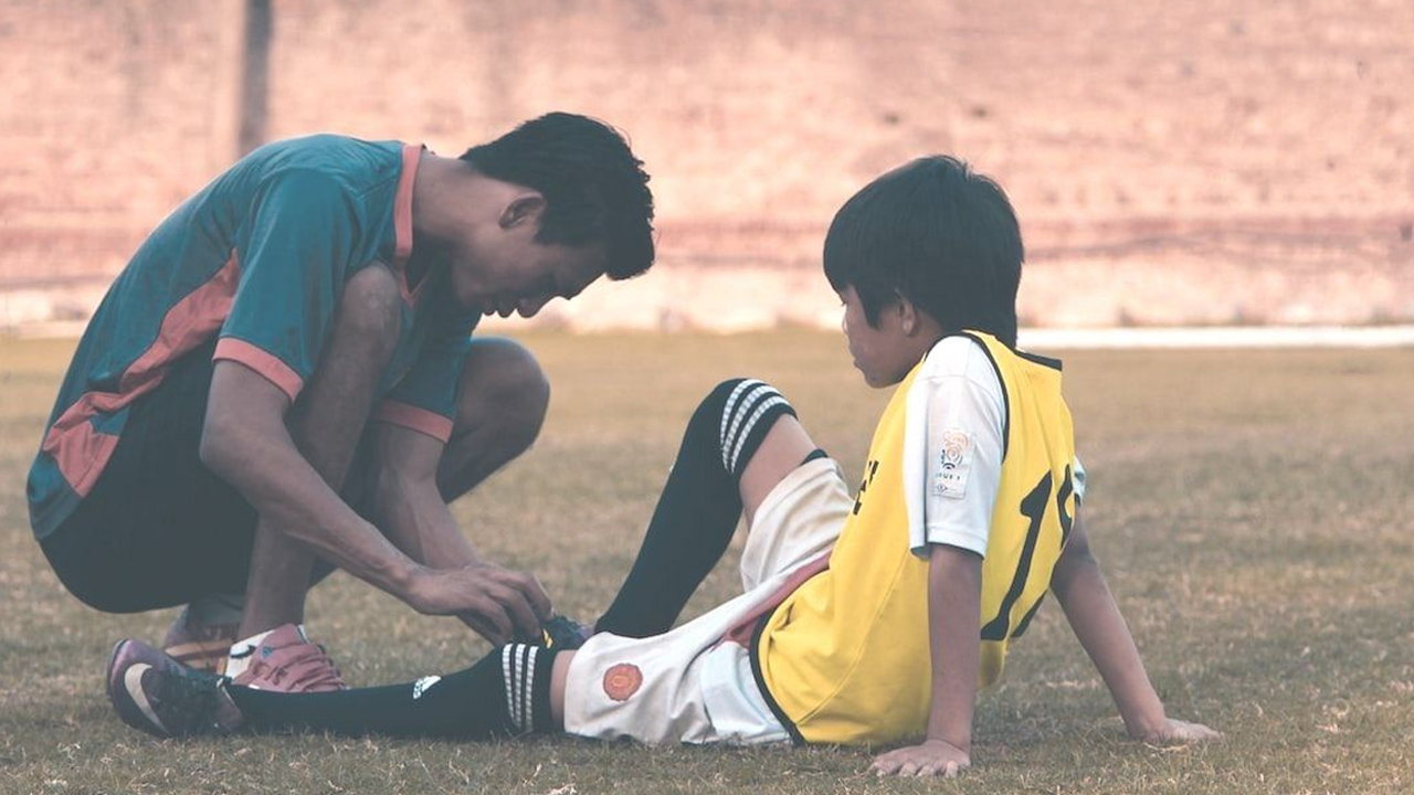 A youth football coach helping a young player on the ground by checking or tying his boot during a match.