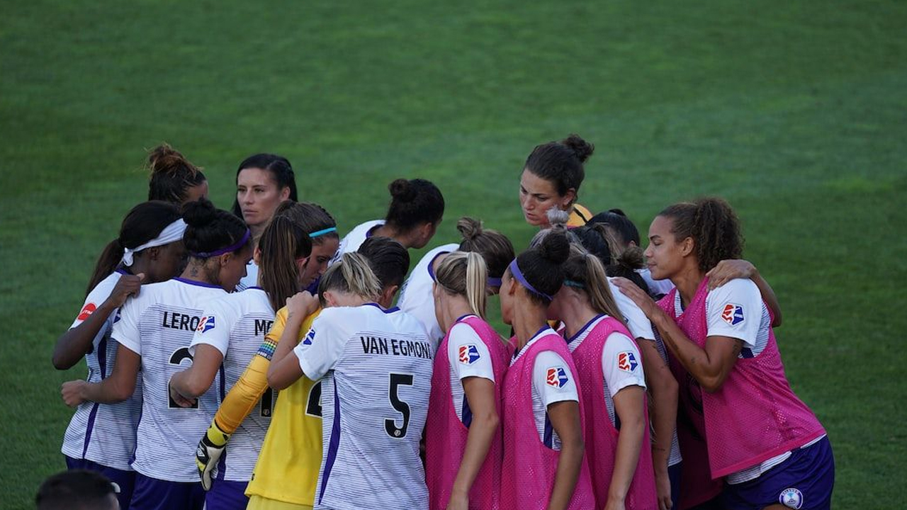 A women’s football team standing in a tight huddle on the pitch before the match begins.