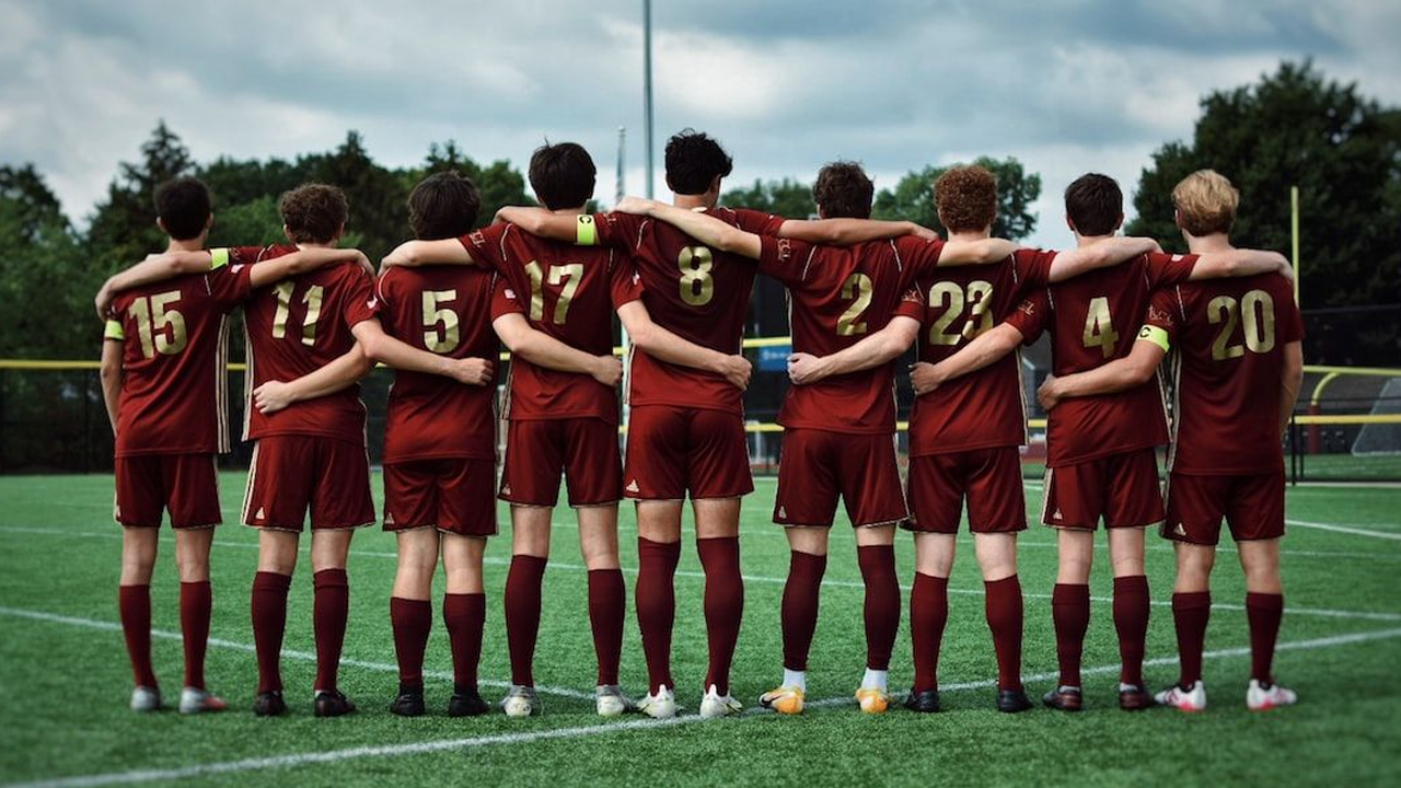 A youth football team standing side by side with their arms linked, wearing matching maroon kits and facing the pitch as they prepare together before a match.