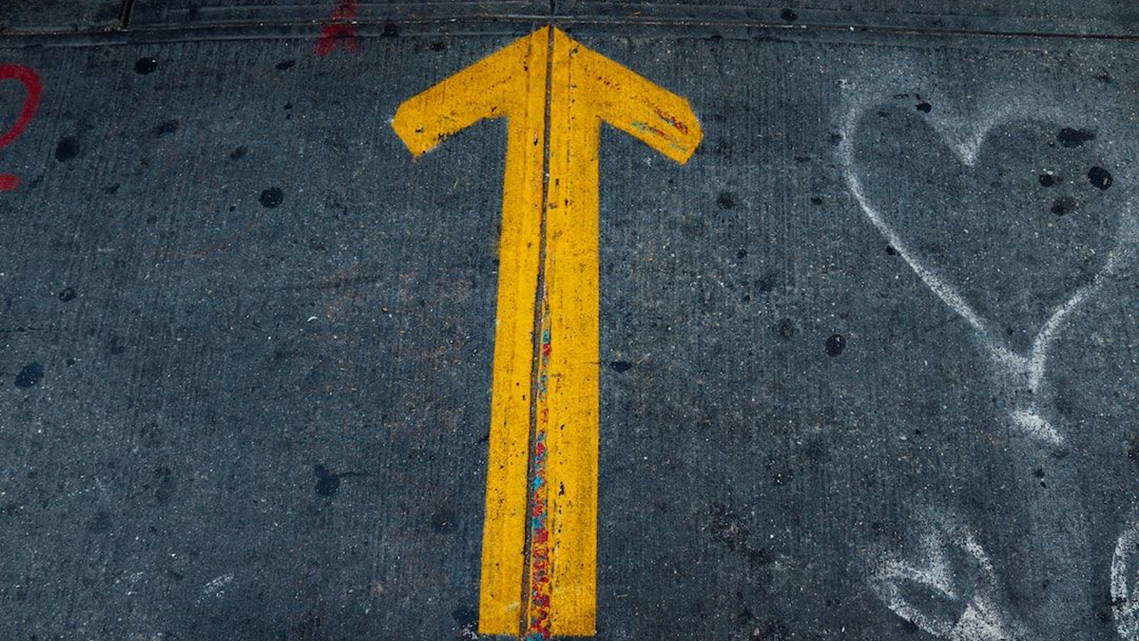 A yellow arrow painted on a concrete pavement pointing forwards, with a chalk-drawn heart visible to the right.