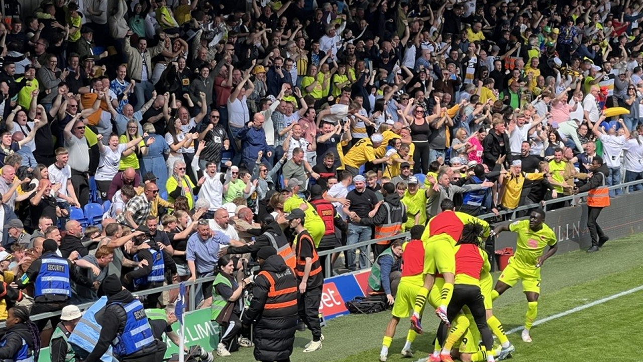 A packed stand of Port Vale supporters celebrating wildly during a match, with stewards positioned at the front. Players in bright yellow kits are gathered near the crowd, some jumping on teammates in a goal celebration.
