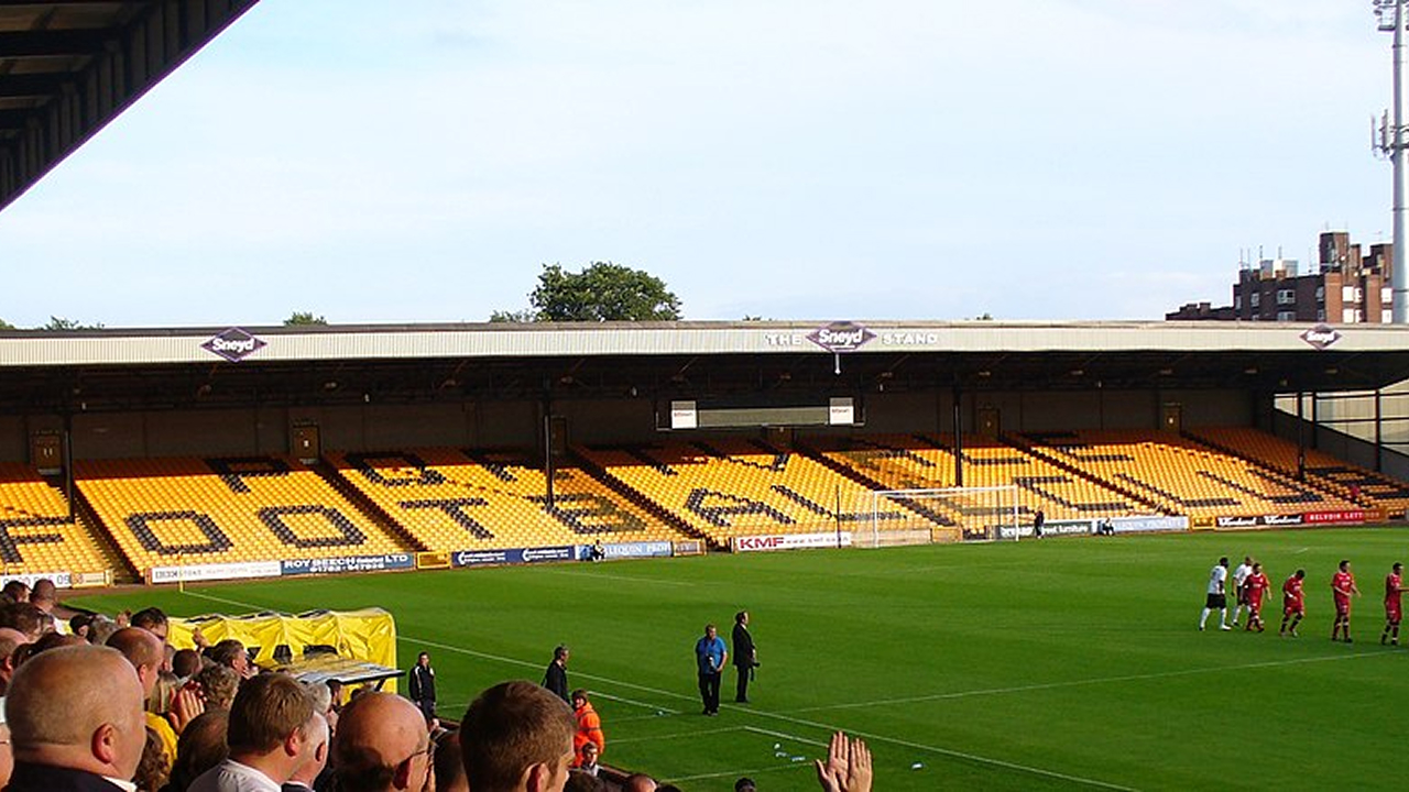 A view of a Port Vale FC home match at Vale Park, showing the large stand with yellow seating spelling out “PORT VALE” and “FOOTBALL CLUB”. Supporters are gathered in the foreground, while players and staff stand on the pitch near the touchline.