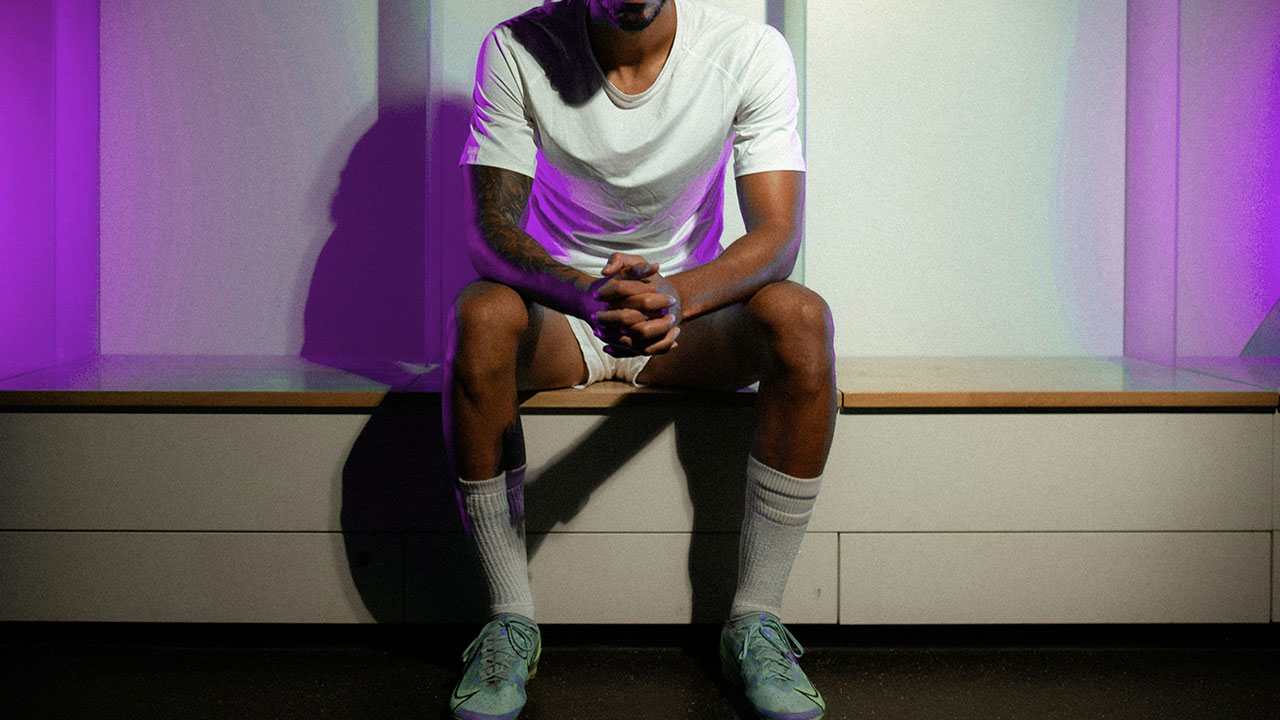 A footballer sitting alone in a changing room, hands clasped, head bowed, lit by a purple light.