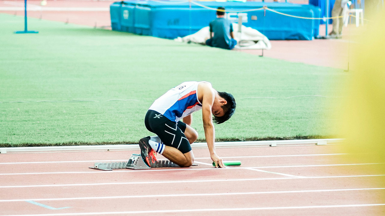 A relay runner crouched at the starting blocks, holding a baton, preparing to race on an athletics track.