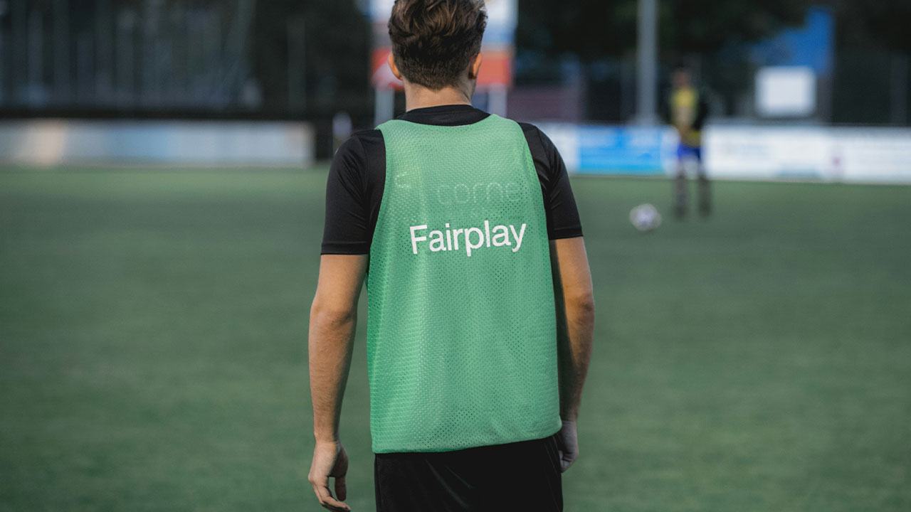 A footballer in a green training bib standing on a pitch, back to the camera, watching a teammate in the distance.