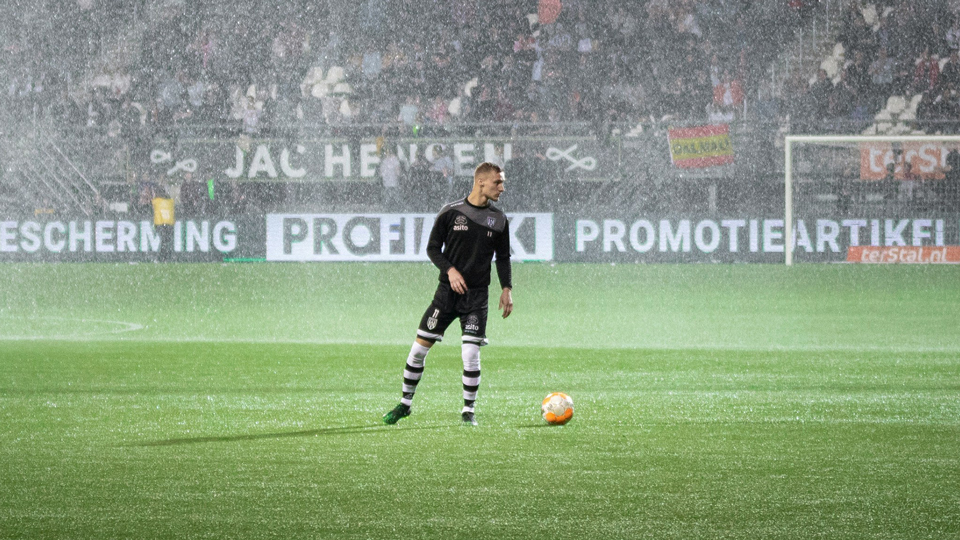 Footballer in a black kit standing over an orange ball on a floodlit pitch during heavy rain, with a packed stadium in the background.