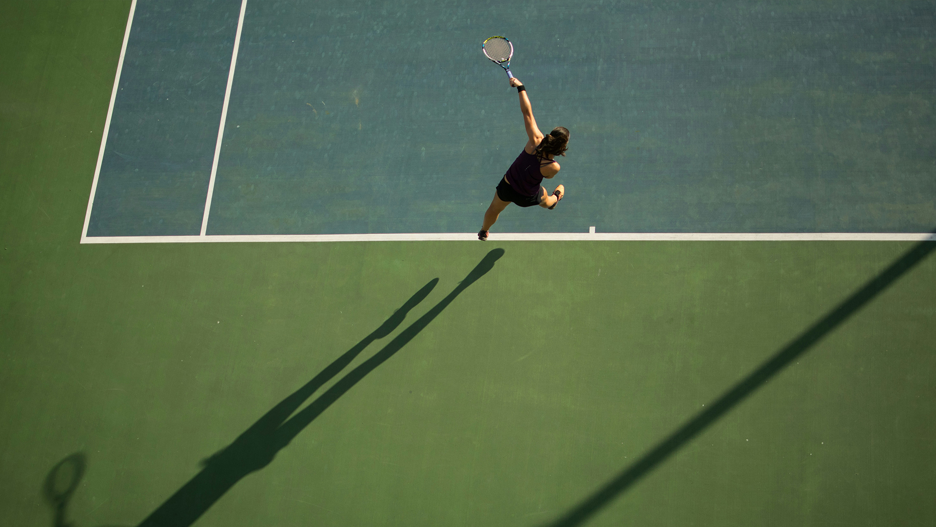 Overhead view of a tennis player jumping to serve on an outdoor hard court, casting a long shadow across the green surface.