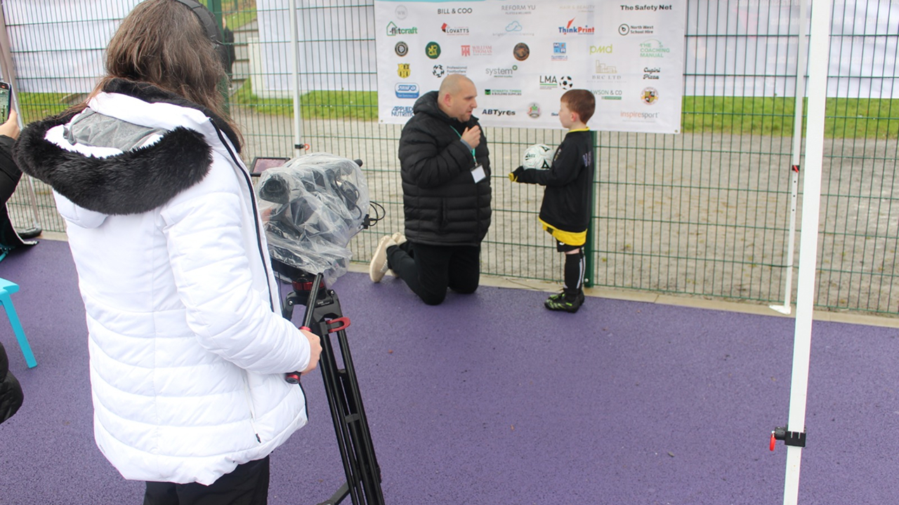 A young grassroots football player being interviewed on camera at the Coachathon, with sponsor boards displayed behind him.