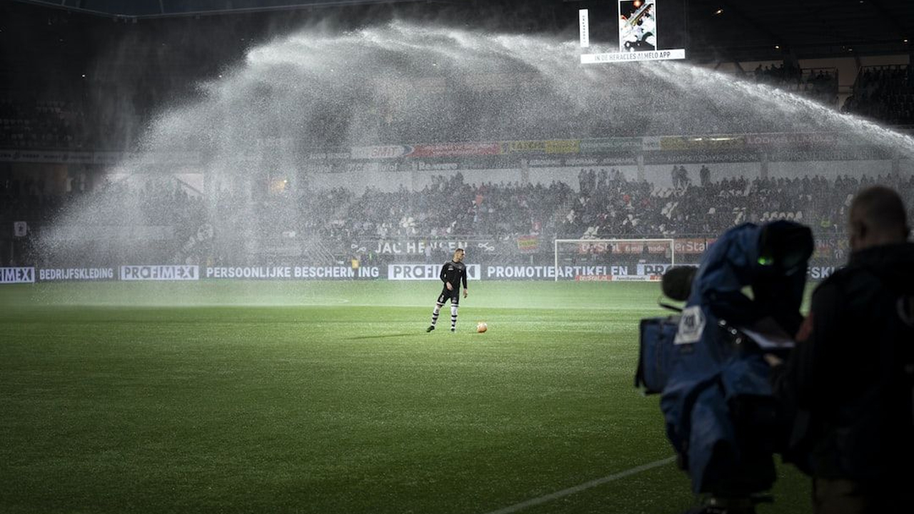 A footballer standing alone on the pitch during a stadium sprinkler spray, with a broadcast camera filming from the sideline and a full crowd in the background.