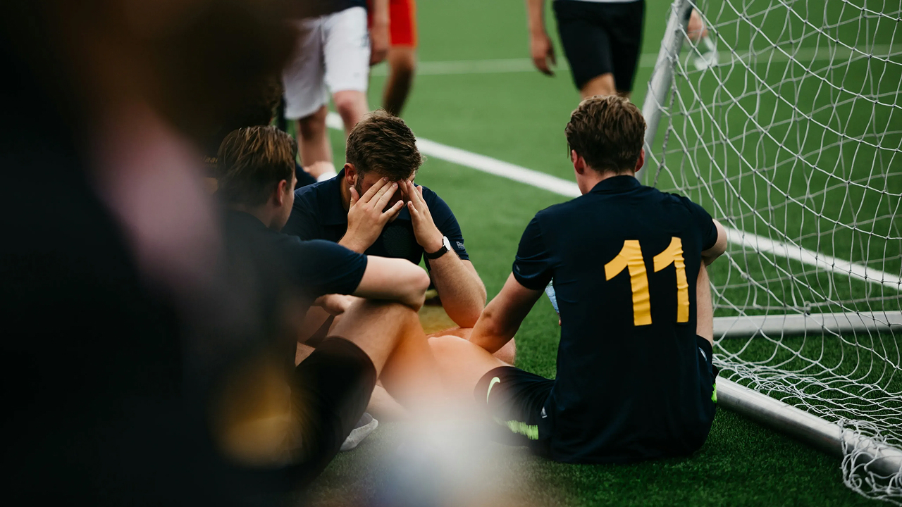 A young footballer sitting on the pitch with his head in his hands, while teammates sit nearby after a match.