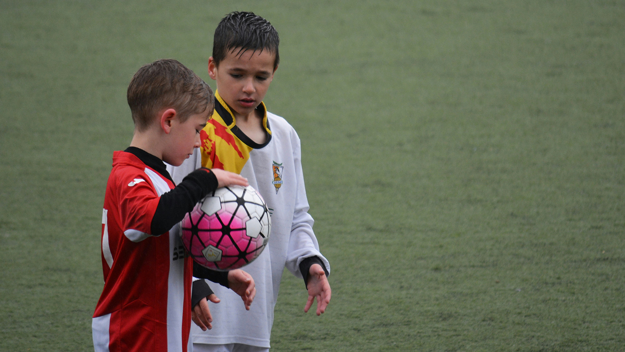 Two young footballers standing on a pitch, one holding a football while they talk.