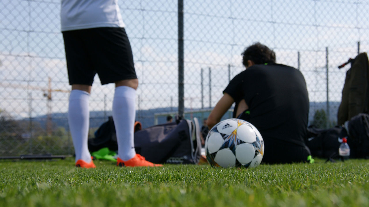 A football resting on the grass beside players sitting and standing near the edge of a pitch.