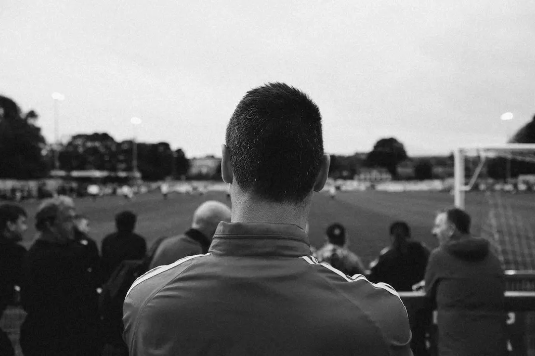 Black and white photo of a person from behind watching a football match from the sidelines, surrounded by other spectators, with a football field and goalpost in the background.